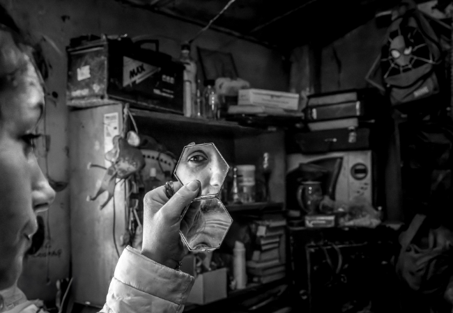 Black-and-white photograph of a hand holding mirror fragments reflecting an eye in a workshop. Copyright Nicolas Wieërs and David Vannussi.