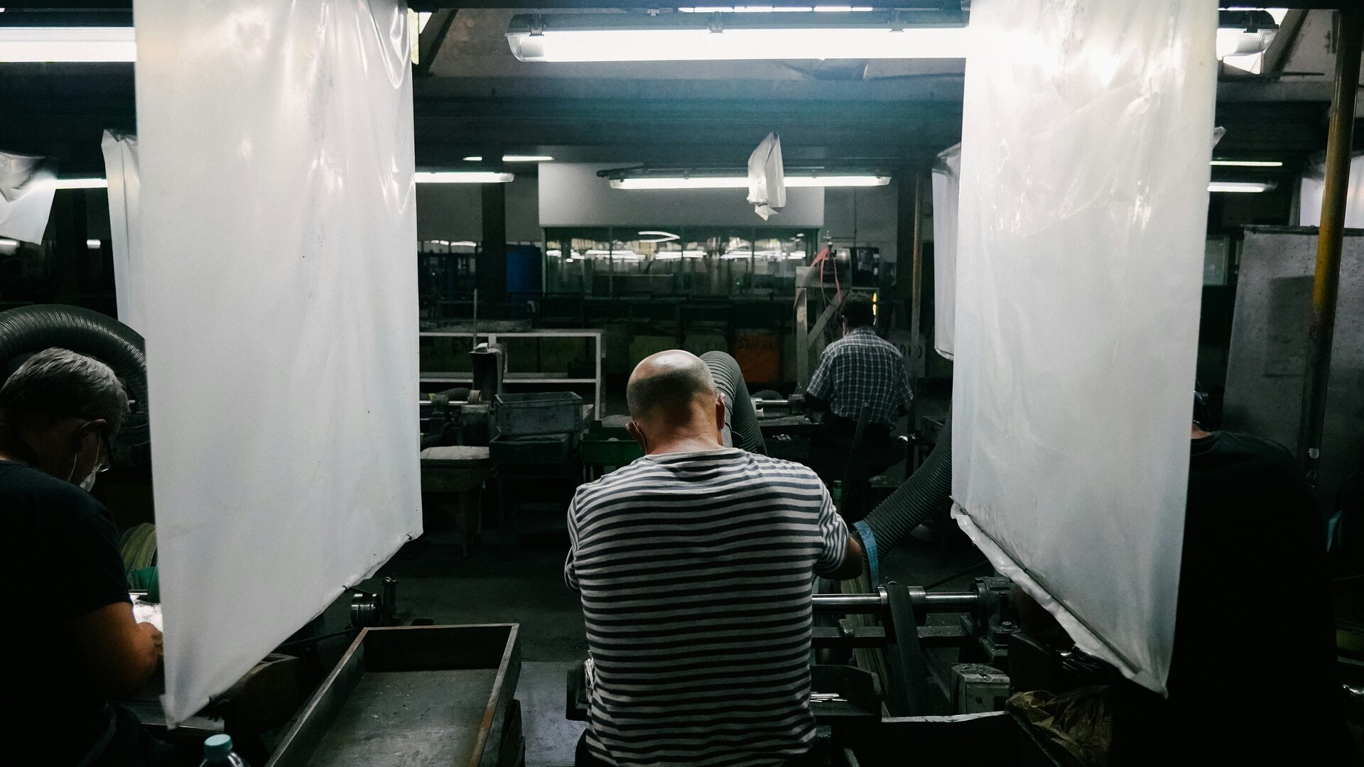 Dim factory with workers at machines; a man in a striped shirt sits in the foreground.