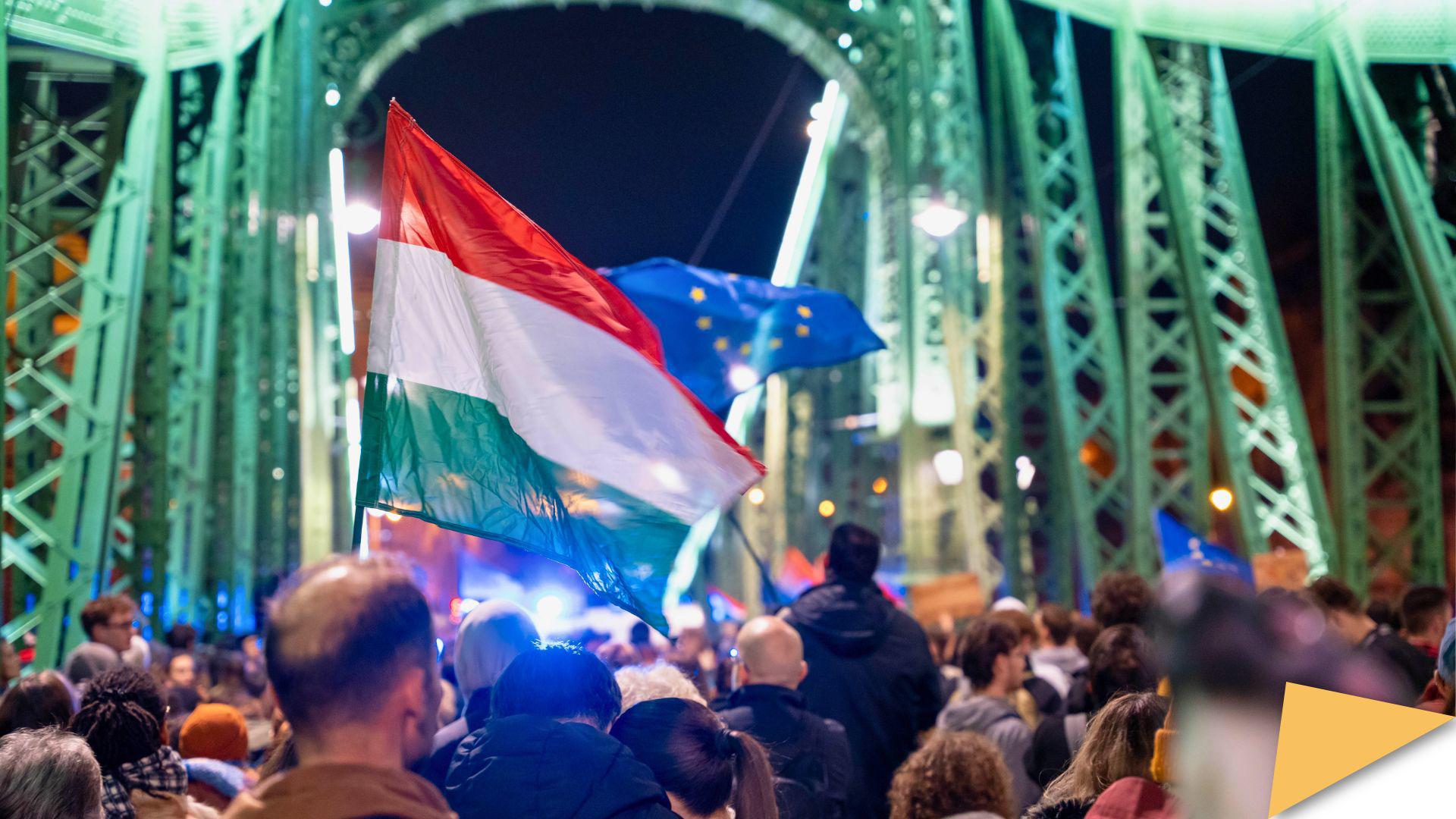 Crowd gathered on a bridge in Budapest, with Hungarian and EU flags visible at night