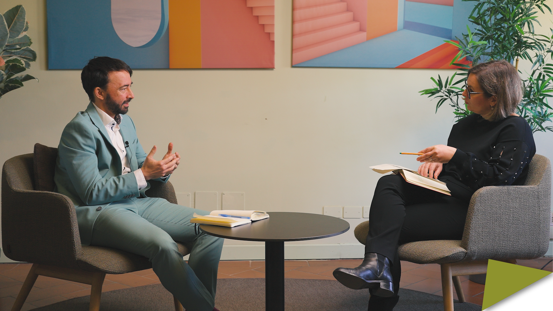 Two researchers in armchairs talk during a recorded discussion, with a table and books between them.