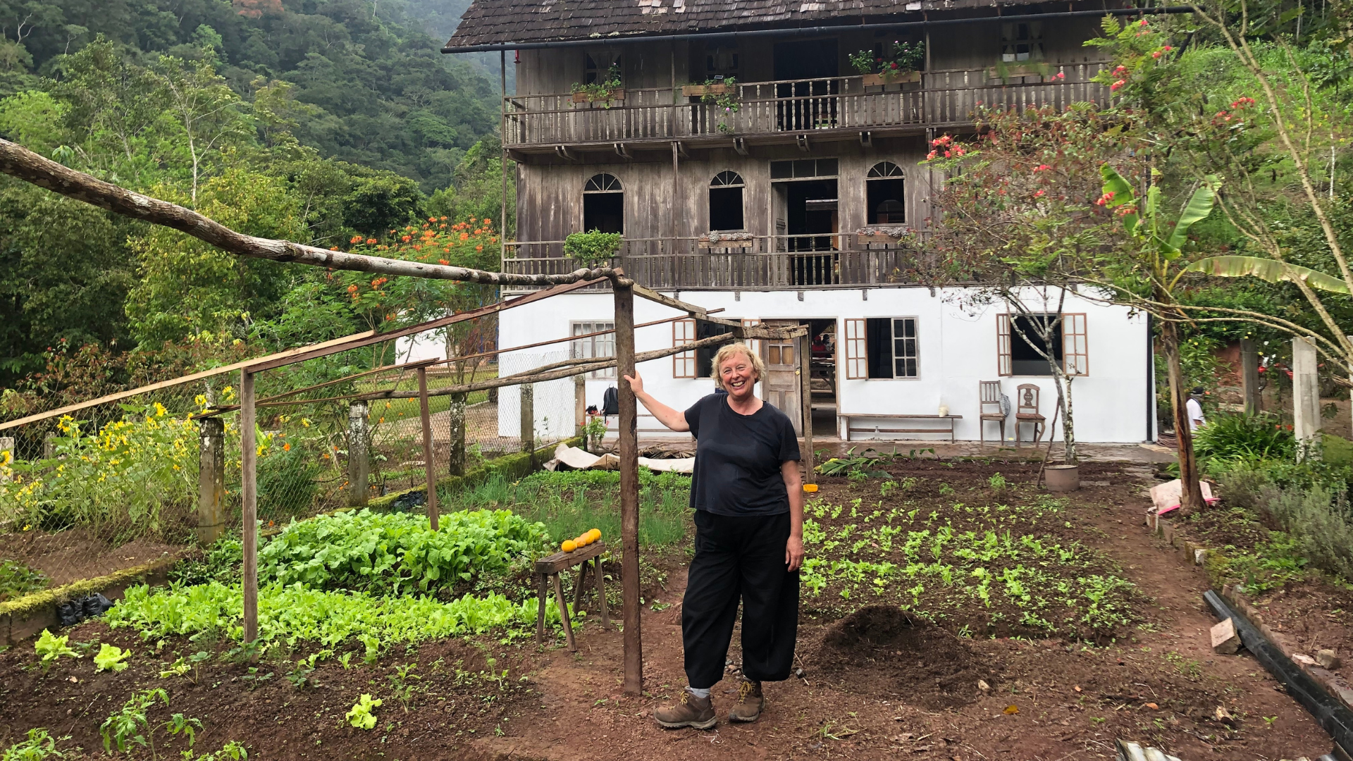 A woman stands smiling in a vegetable garden in front of a tall wooden house with balconies and arched windows, backed by forested hills.
