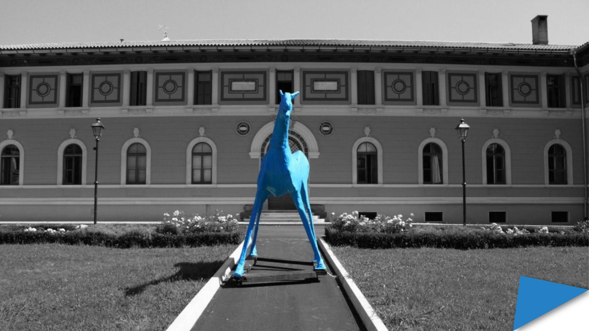 Marco Cavallo, a large blue horse sculpture, stands on a path in front of the former psychiatric hospital in Trieste. The building behind it is in black and white.