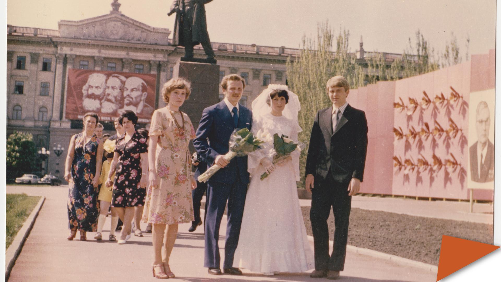 Bride and groom with guests posing in a Soviet-era city square, with Marx, Engels, and Lenin banner and statue in the background.