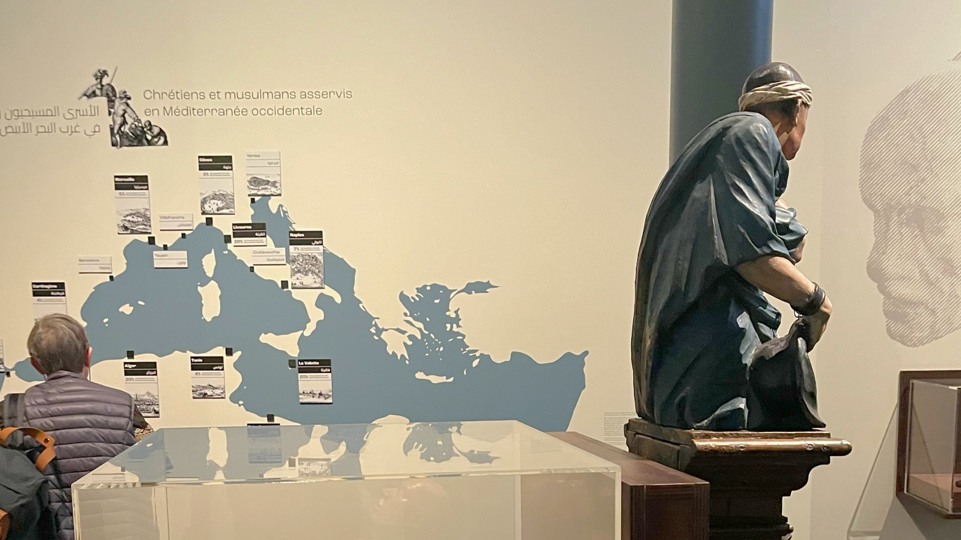 A sculpture of a kneeling, bound enslaved man before a large wall map of the Mediterranean, with a visitor seated nearby.