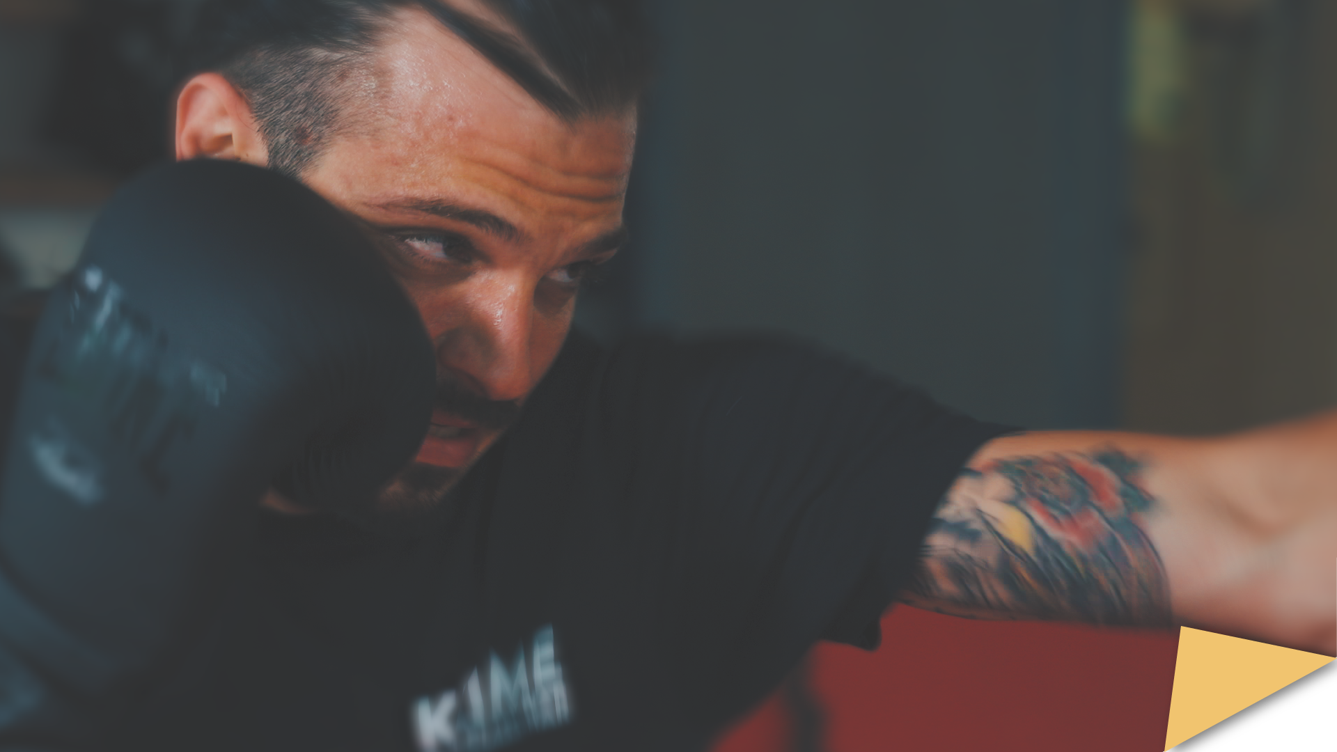 Close-up of a man boxing, wearing black gloves and a T-shirt, with a visible tattoo on his arm.