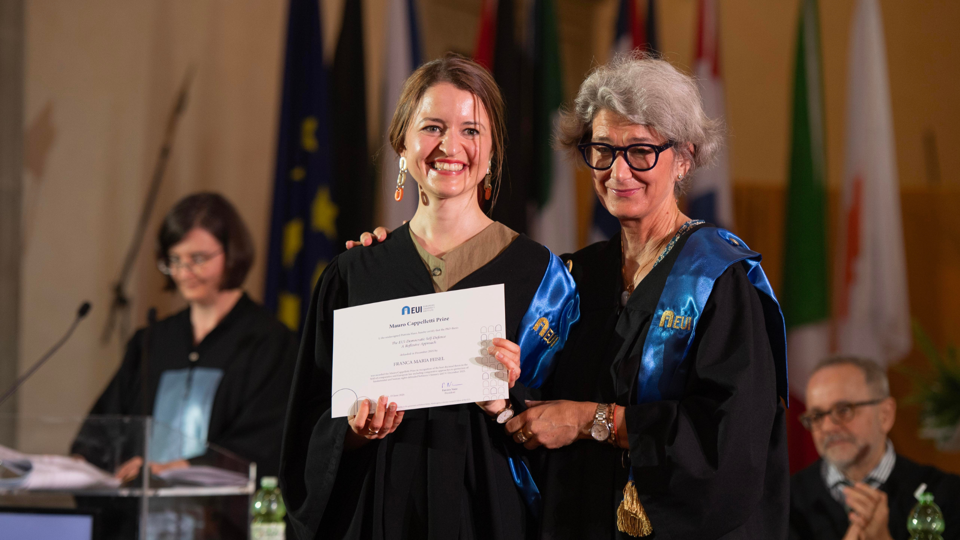 Franca Maria Feisel and EUI President hold her Mauro Cappelletti Prize certificate together at the Conferring Ceremony, in front of a row of flags.