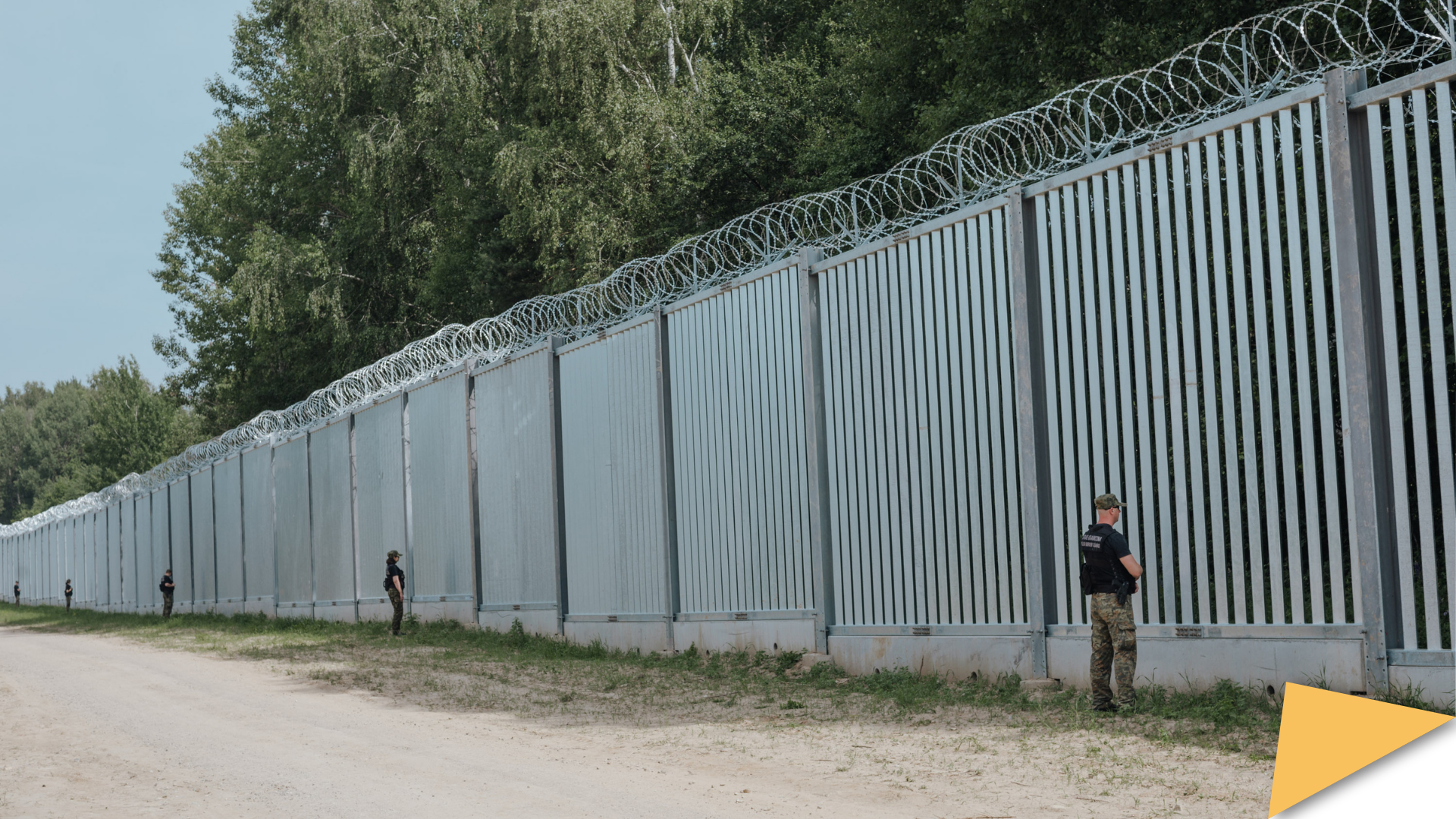 Metal border barrier topped with razor wire along a forested area, with uniformed guards standing at intervals.