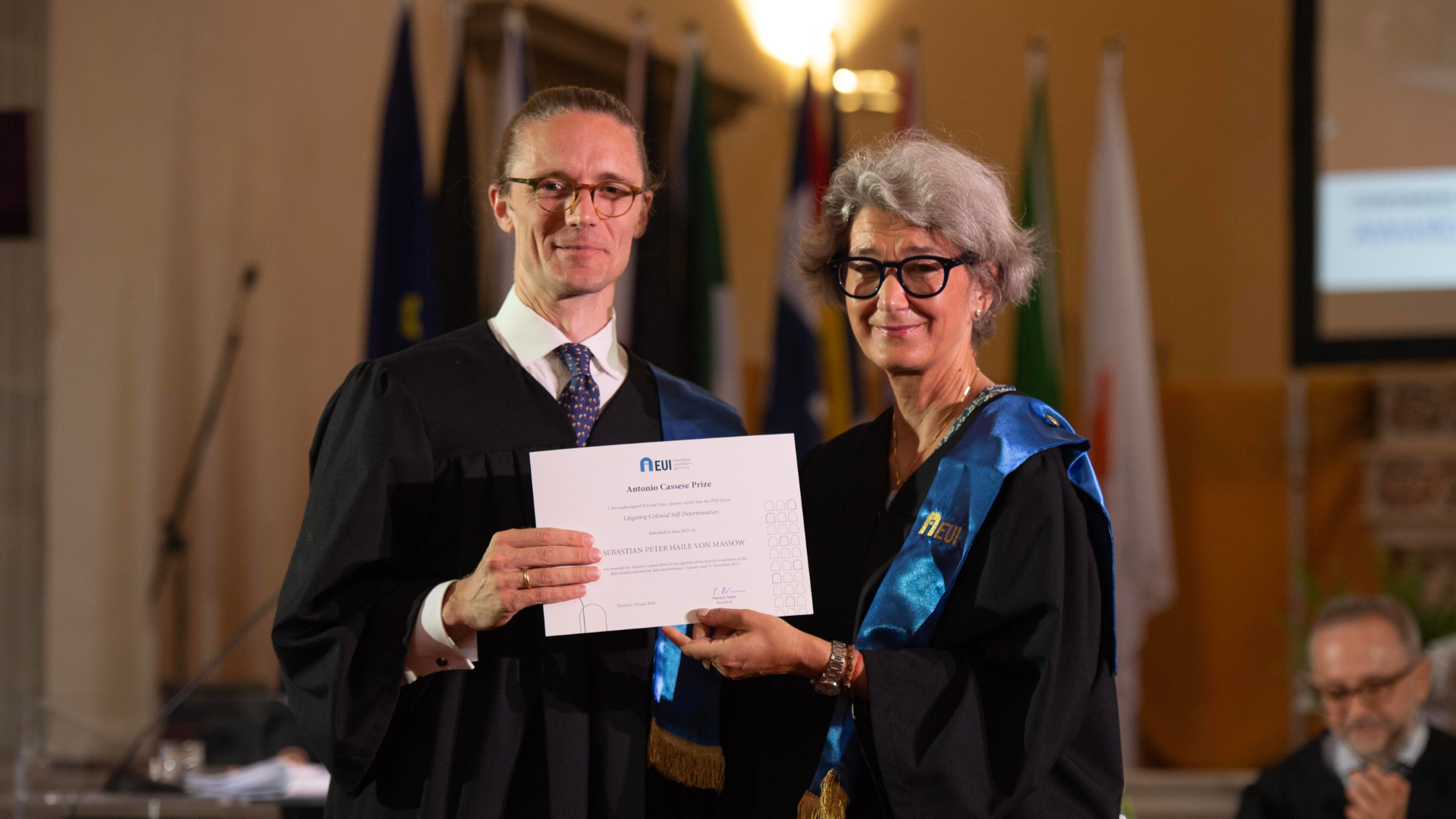 Sebastian von Massow and EUI Presidenthold his Antonio Cassese Prize certificate together at the Conferring Ceremony, in front of a row of flags.