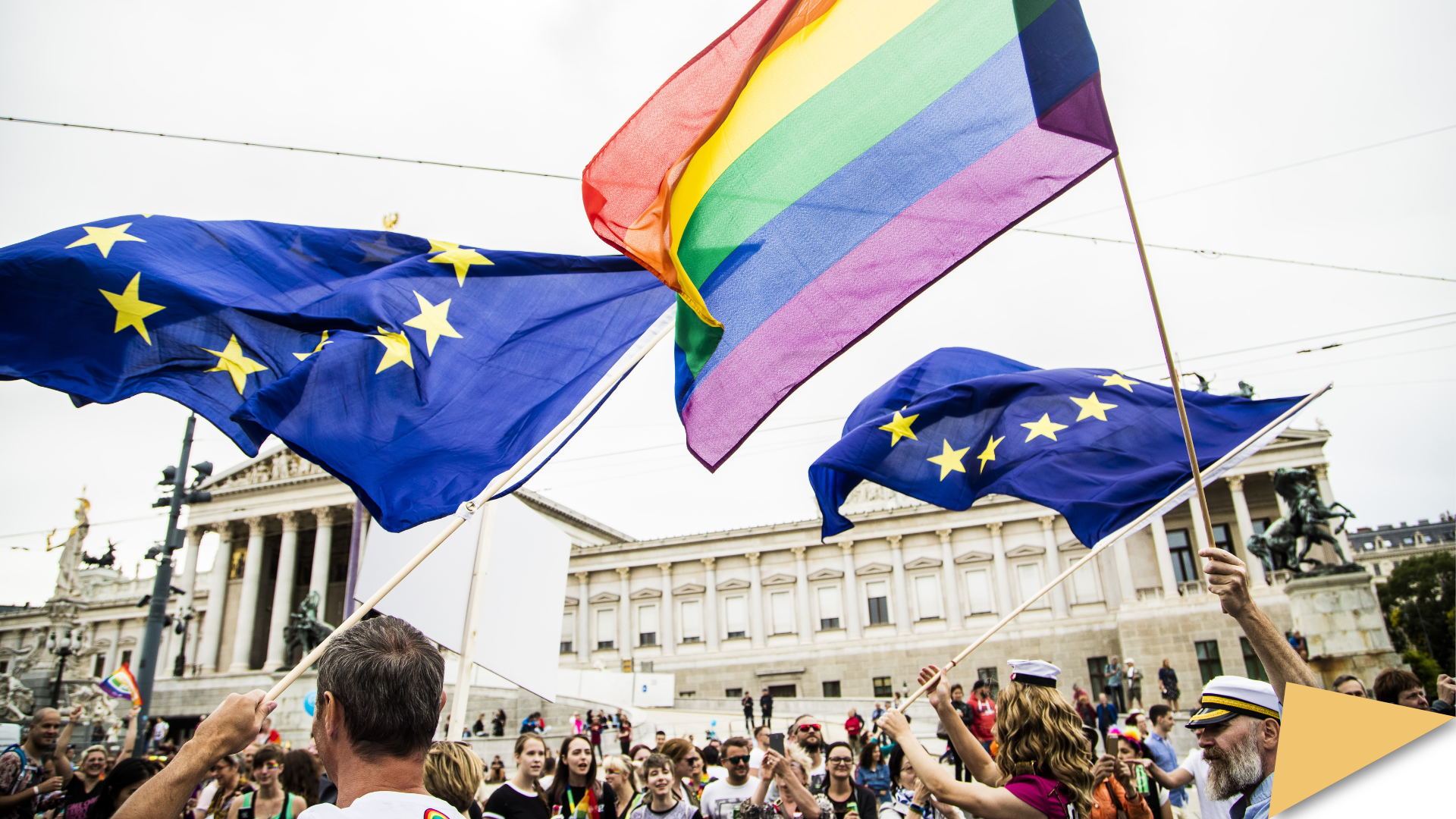 Rainbow Pride flags and EU flags wave together above a crowd at a Pride march outside the Austrian Parliament in Vienna.