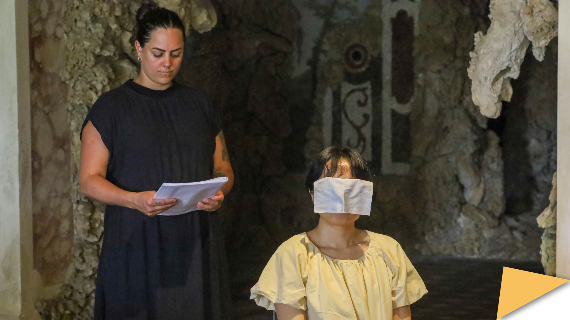A standing summer school participant reads while another sits blindfolded in a reenactment scene.