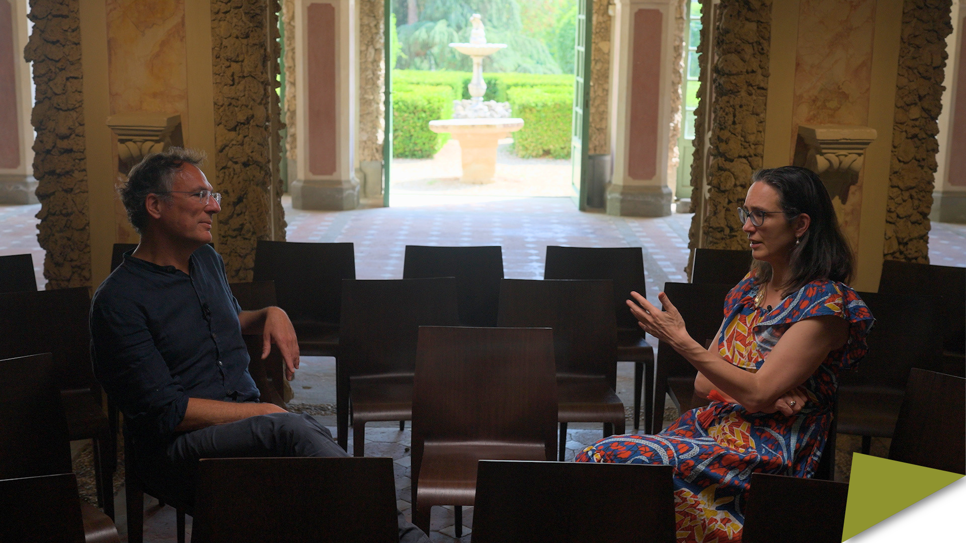 Arnulf Becker Lorca and Sarah Nouwen in conversation on wooden chairs inside the EUI Grotto, a garden fountain framed in the doorway behind them.