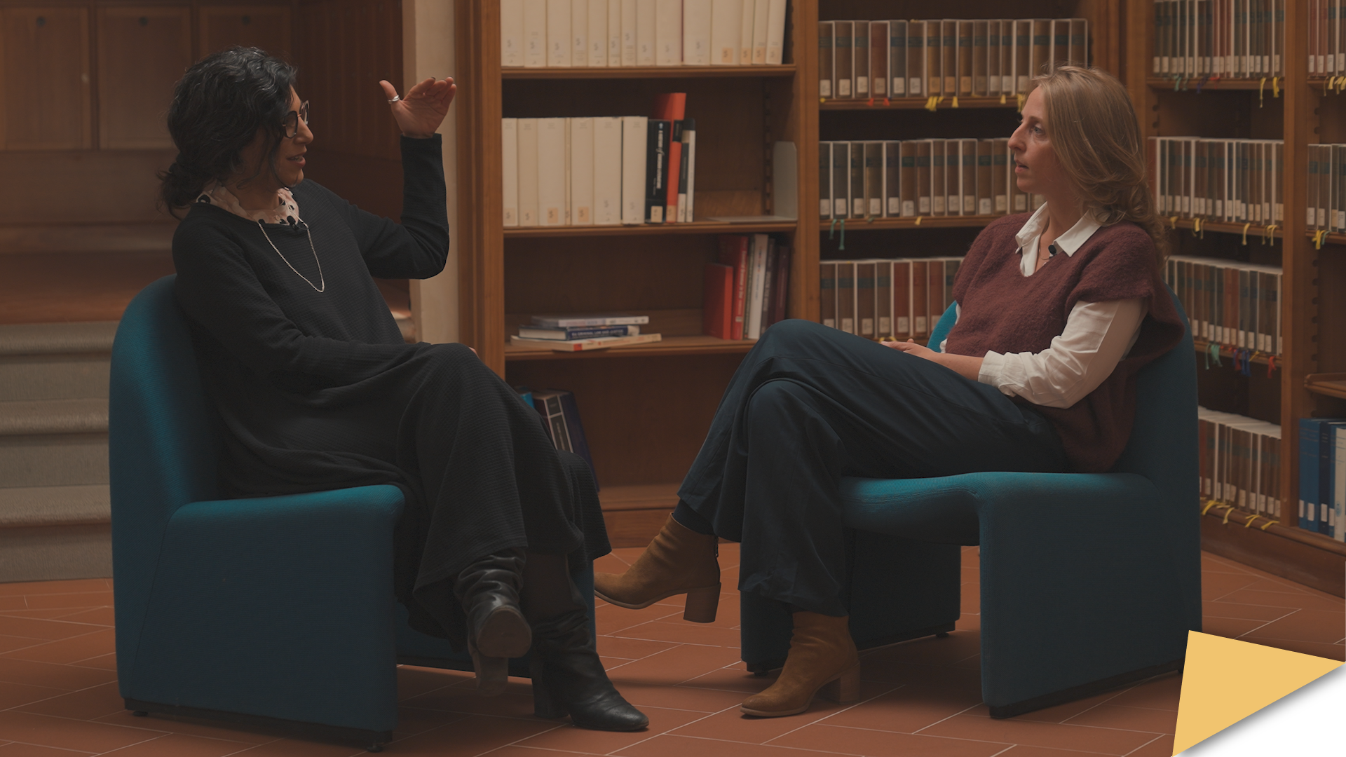 EUI scholars Revital Madar and Caitlin Procter sit facing each other in blue chairs, engaged in a conversation for the EUI UpFront video. They are in a library setting, surrounded by bookshelves.