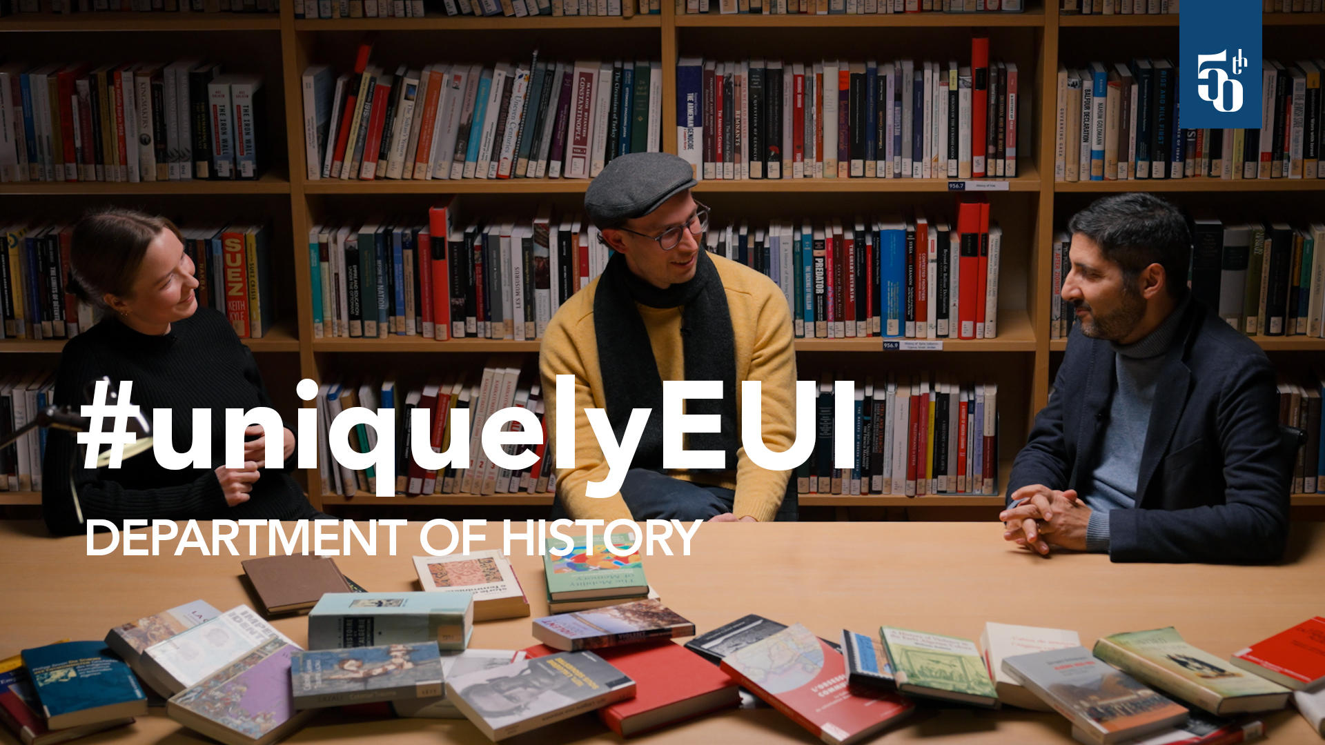 Three researchers sit around a table covered with books in a library, in conversation, marking the History Department’s anniversary video.