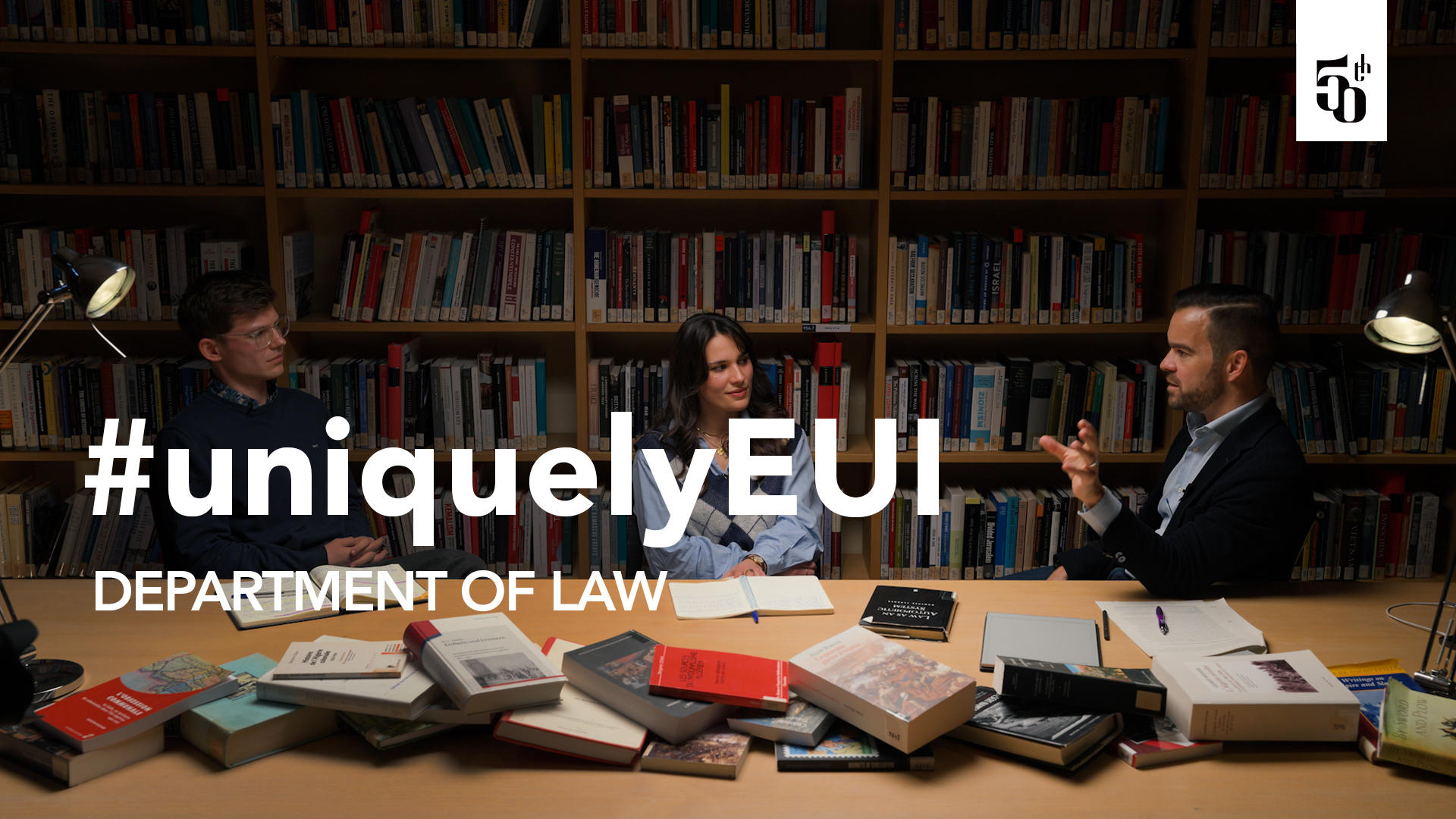 Three scholars sit at a table in front of bookshelves at the EUI Library, speaking during a filmed discussion for the #uniquelyEUI Department of Law anniversary video.